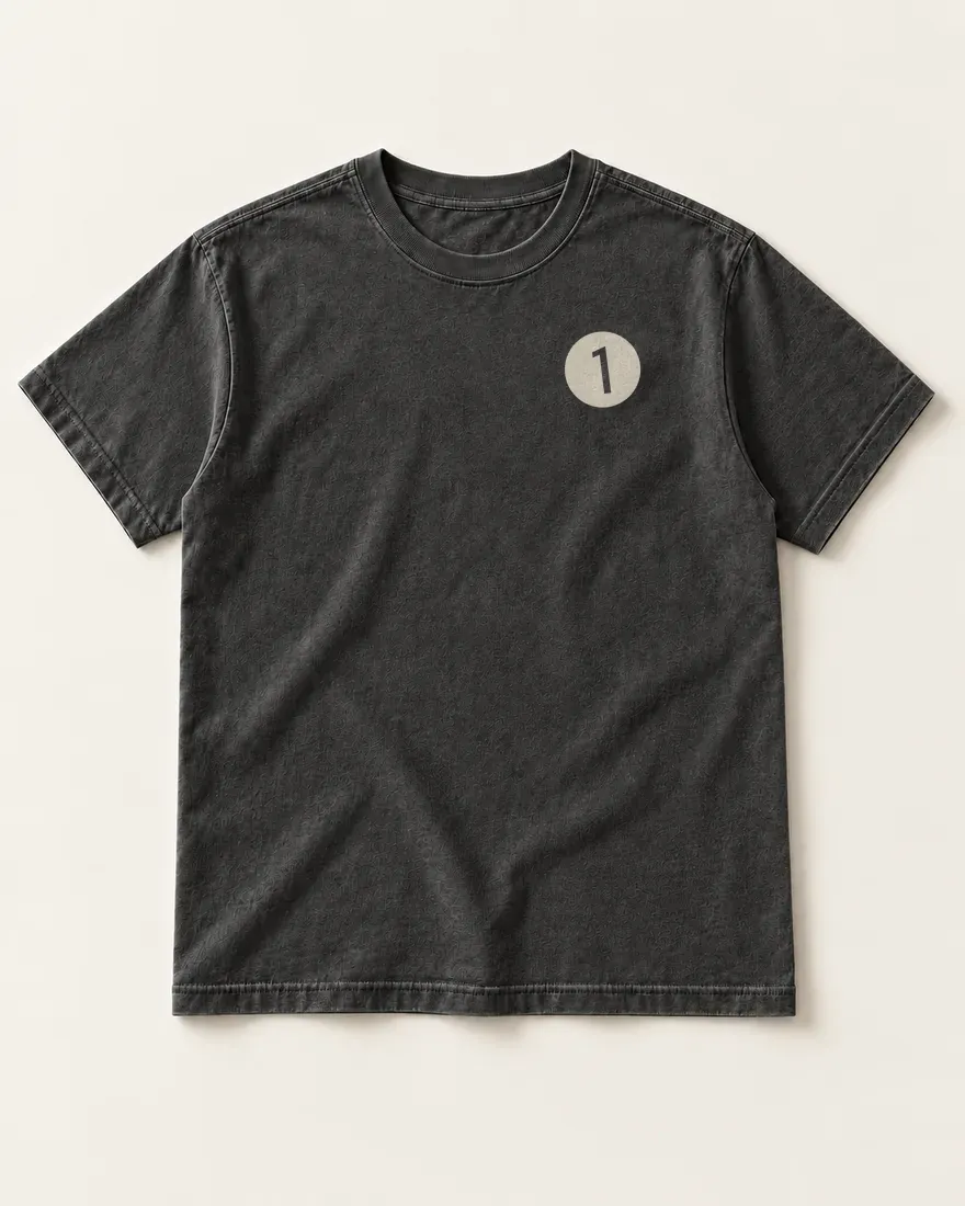 The front of a washed black tee laid flat: a small cream disc on the left chest, the 1 inside it left in the shirt’s own black.