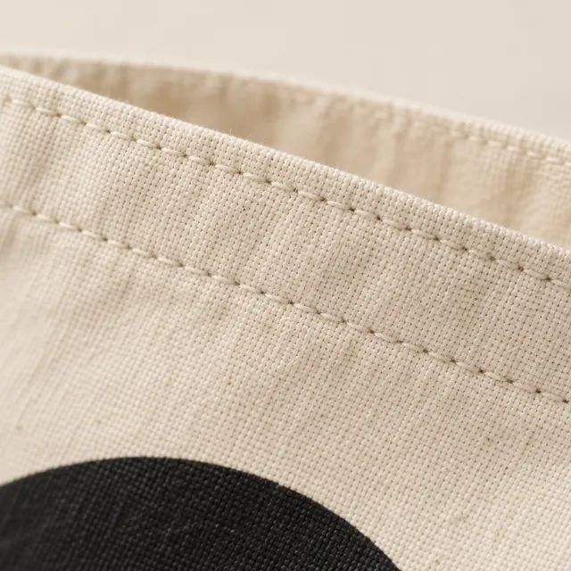 The folded top hem of a canvas tote, two rows of stitching, the edge of a black disc below.
