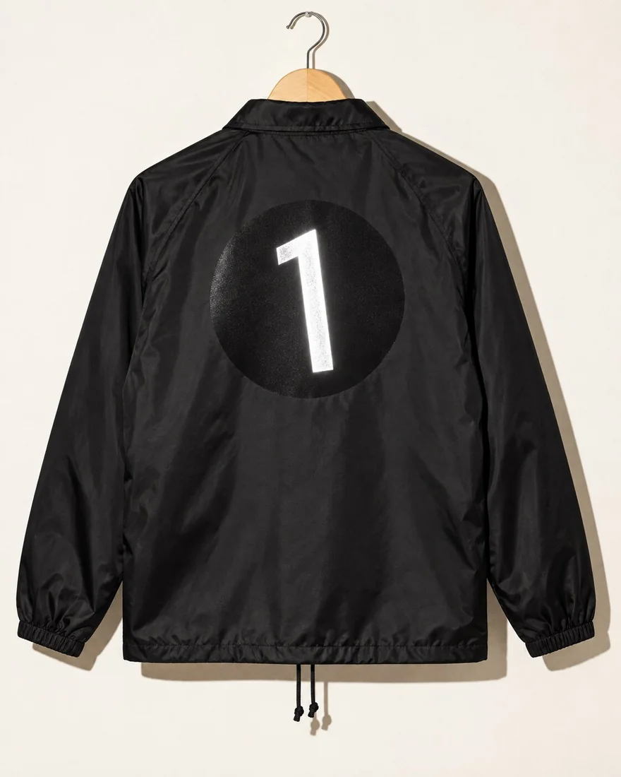 In a camera flash: the back of a black coach jacket on a hanger, the black disc barely showing, its leaning 1 glowing silver.