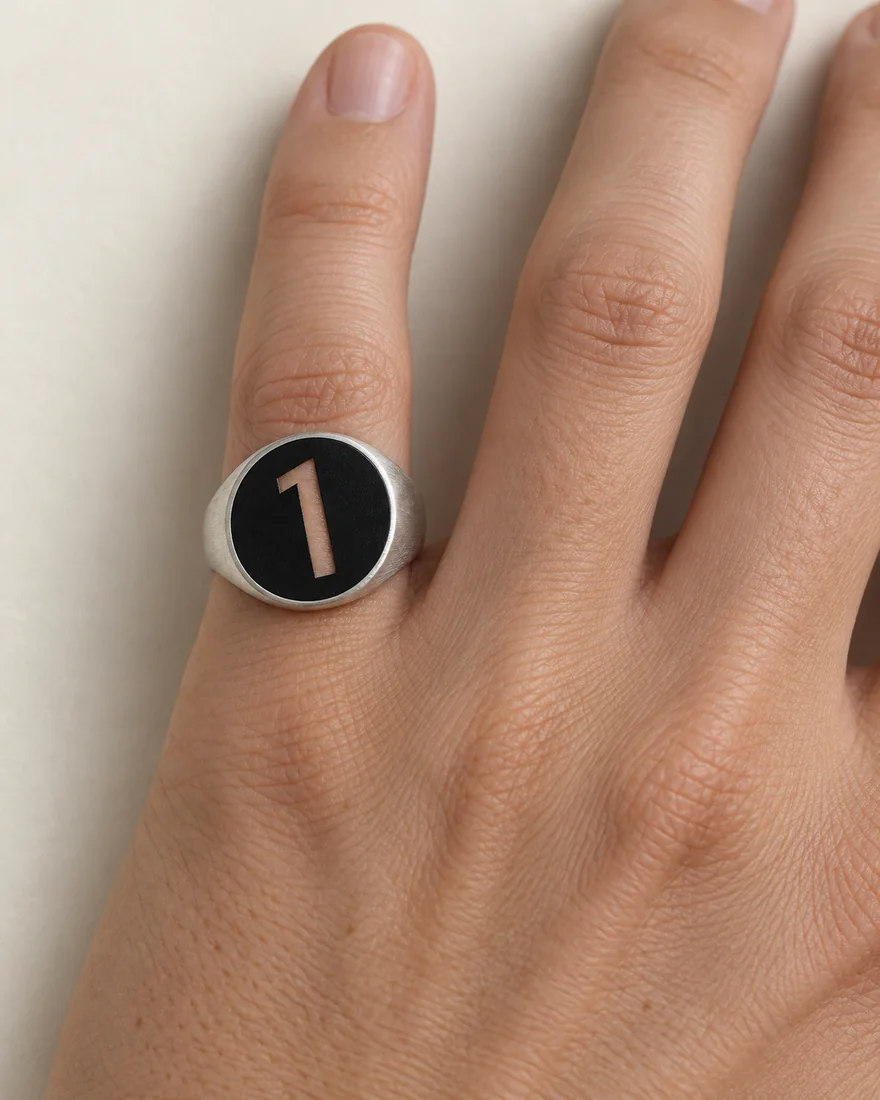 A brushed silver signet ring on a hand, its round black face the disc, the leaning 1 cut through so the skin shows as the 1.